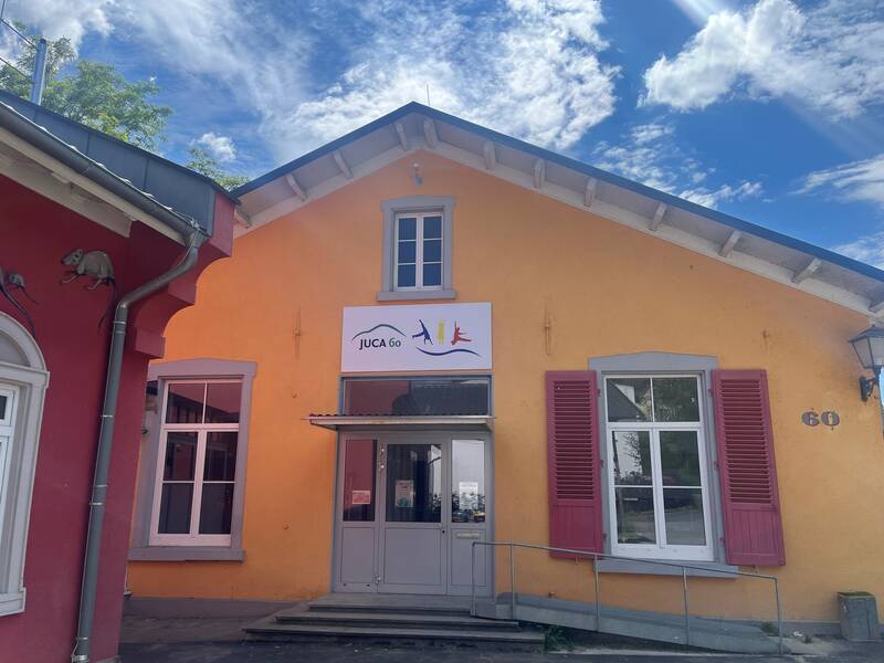Ein gelbes Gebäude mit einem großen Fenster und roten Fensterläden hat ein Schild mit der Aufschrift "JUCA 60" über dem Eingang.