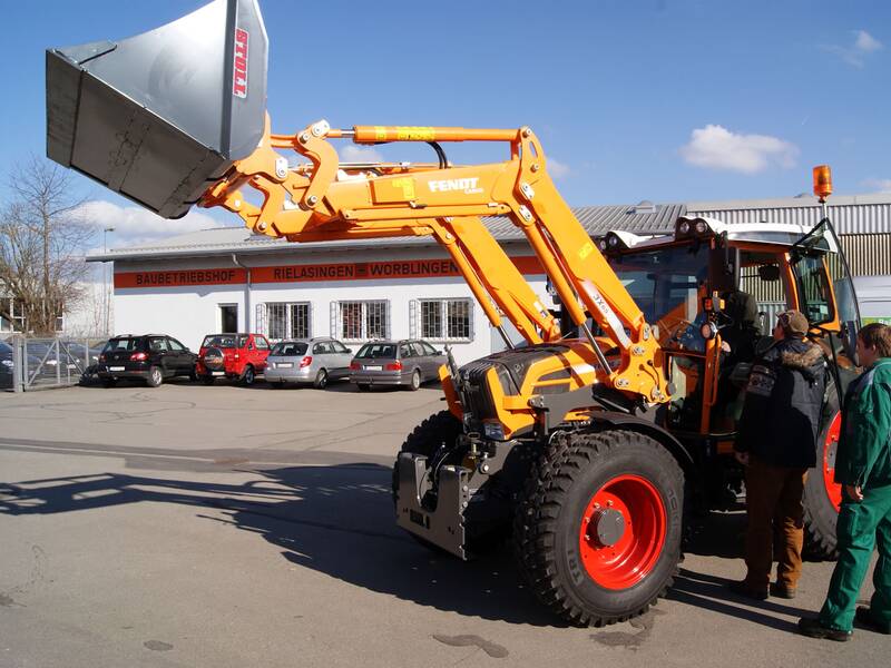 Ein leuchtend orangefarbener Traktor mit einem Frontlader steht auf einem Parkplatz, umgeben von drei Menschen, während im Hintergrund ein Gebäude mit Fahrzeugen sichtbar ist.