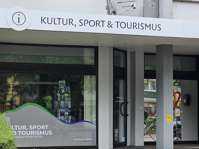Der Eingang eines Gebäudes mit einem Schild "Kultur, Sport & Tourismus" und großen Fenstern, die eine Inneneinrichtung mit Broschüren zeigen.