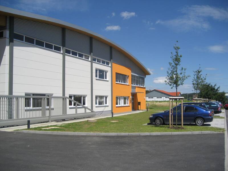 Ein modernes Gebäude mit einer Kombination aus weißen und orangefarbenen Wänden steht neben einem Parkplatz und einer Wiese, auf der junge Bäume wachsen.