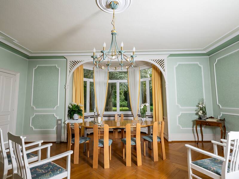 Ein elegantes Trauzimmer mit hellgrünen Wänden, Parkettboden, einem großen Tisch mit Stühlen in der Mitte und einer großen Fensterfront mit weißen Gardinen und gelben Vorhängen.