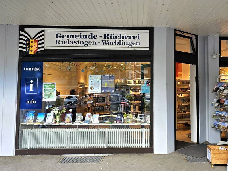 Die Fassade der Gemeindebücherei in Rielasingen-Worblingen mit großen Fenstern, durch die Bücher und Infomaterial sichtbar sind, und einem Eingang auf der rechten Seite.