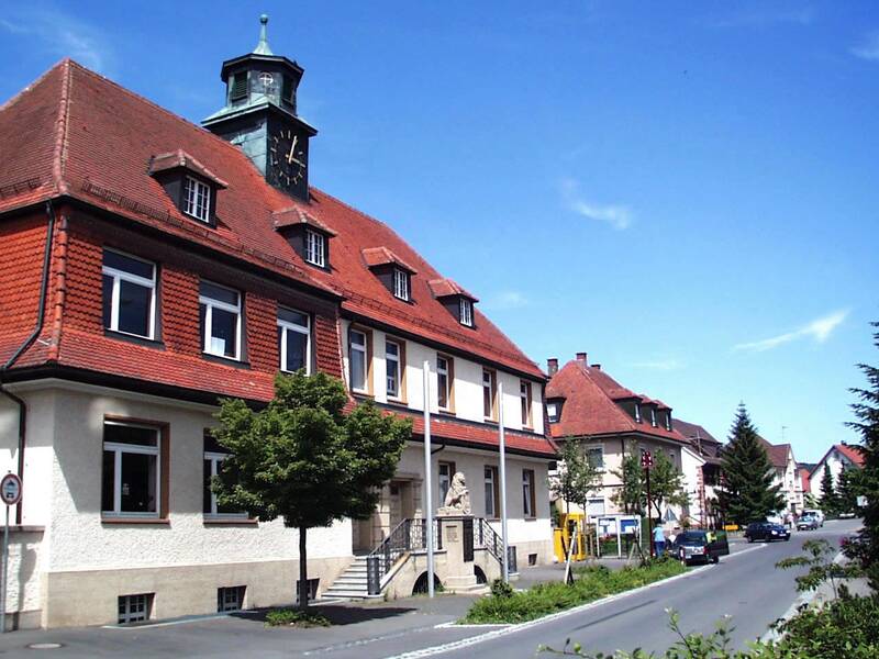 Eine Straße mit mehreren historischen Gebäuden, die rote Ziegeldächer haben, darunter ein größeres Gebäude mit einem Uhrturm.