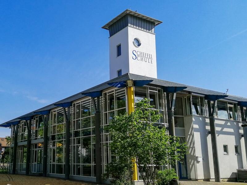 Ein modernes Gebäude mit Glasfassade und einem Turm, auf dem "Scheffelschule" steht, umgeben von blauen Himmel und grünen Bäumen.
