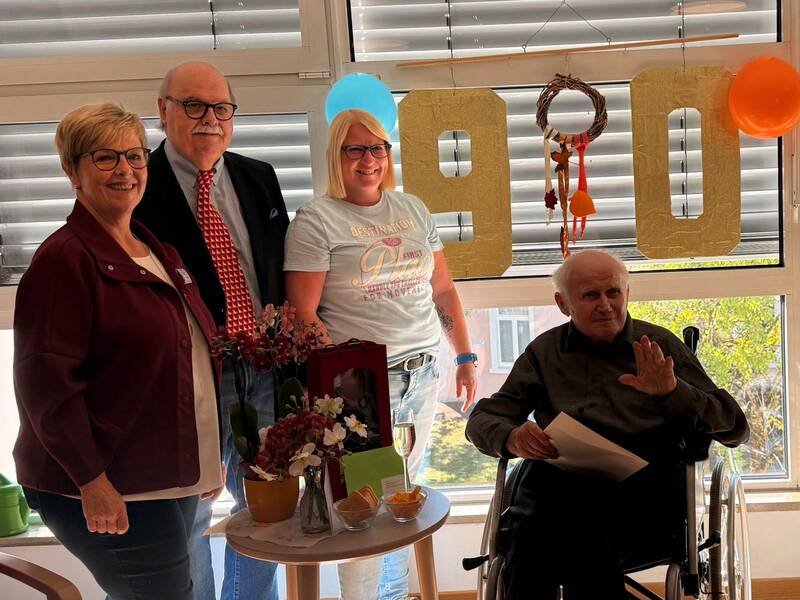 Vier Menschen, darunter eine Person im Rollstuhl, stehen zusammen in einem Raum mit großen goldenen Zahlen "90" im Hintergrund, die auf einen Geburtstag hinweisen.