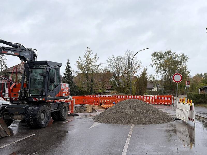 Eine Baustelle mit einem großen Kieshaufen, einem Bagger und Absperrungen blockiert eine Straße in einem Wohngebiet.