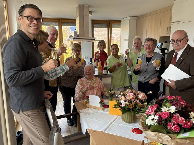 Eine Gruppe von Menschen steht fröhlich in einem Raum, hebt Gläser zum Anstoßen, während eine ältere Frau lächelnd in der Mitte sitzt, umgeben von Blumen.