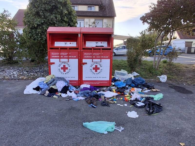 Vor zwei roten Altkleidercontainern des Deutschen Roten Kreuzes liegt eine große Menge unsortierter Kleidung und anderer Gegenstände auf dem Boden.
