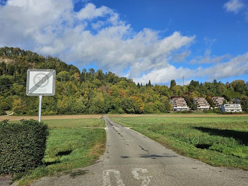 Ein ländlicher Weg führt durch eine grüne Wiese, flankiert von einem aufgehobenen 30er-Zone-Schild, mit Häusern und bewaldeten Hügeln im Hintergrund unter blauem Himmel.