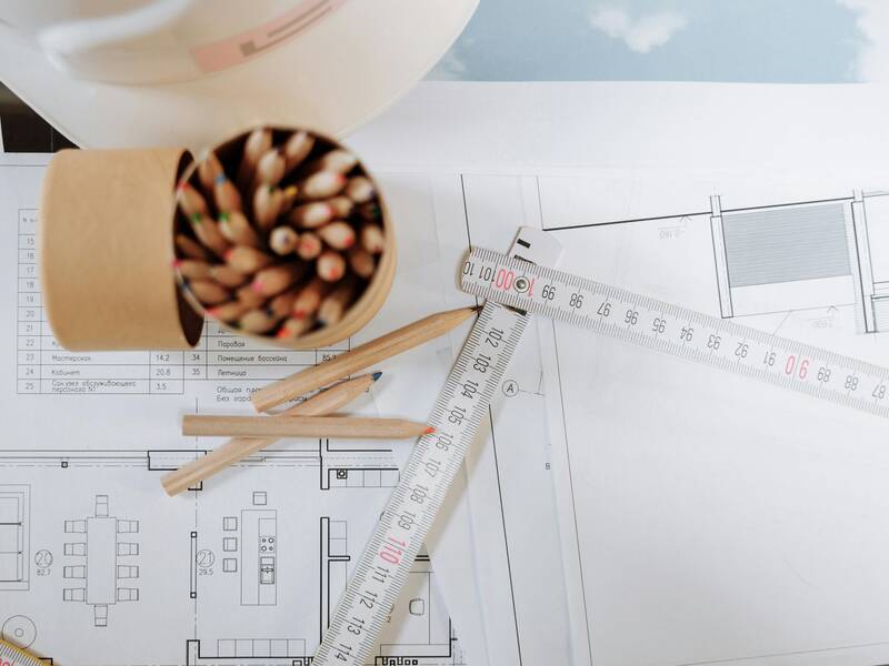 Das Bild zeigt einen Schreibtisch mit Architekturzeichnungen, einem offenen Behälter voller Buntstifte, einem Lineal und einem Schutzhelm.