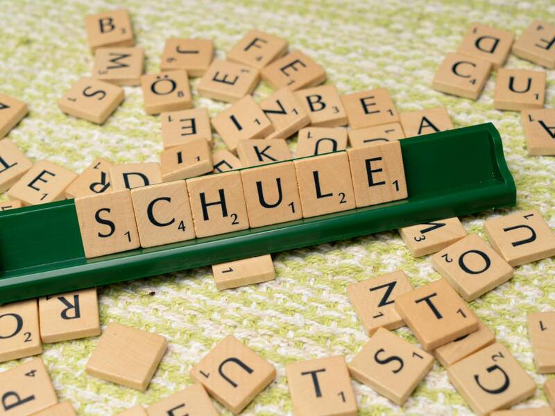 Ein grünes Scrabble-Regal zeigt das Wort "SCHULE", umgeben von verstreuten Buchstaben auf einem stoffartigen Untergrund.