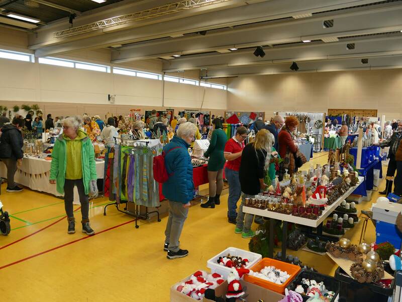 In einer großen Halle betrachten Menschen verschiedene handgefertigte Weihnachtsdekorationen und Geschenkartikel an unterschiedlichen Ständen.
