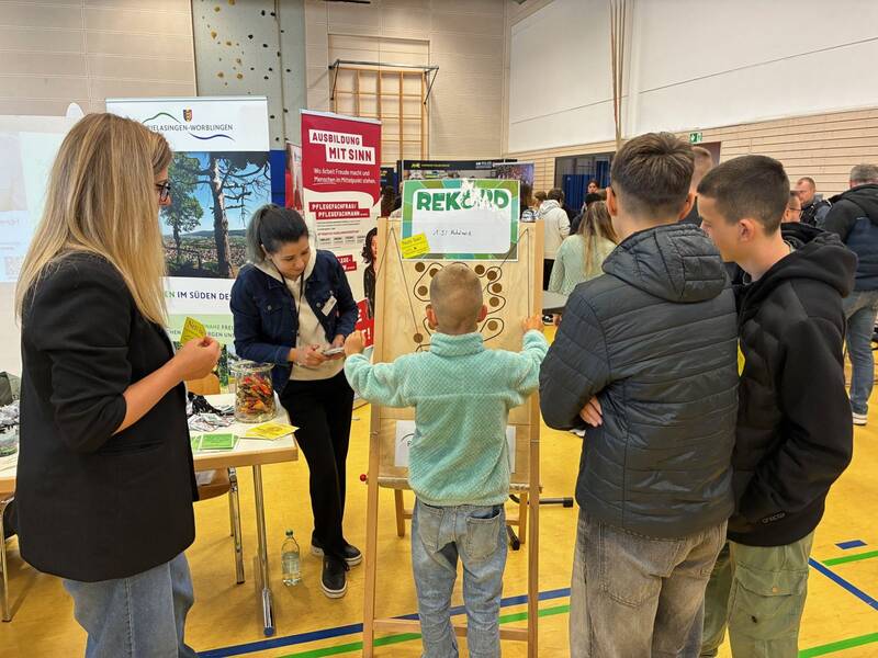 In einer Halle beobachten mehrere Menschen, darunter Kinder und Erwachsene, wie ein Junge ein Spiel auf einer Holztafel spielt, während Informationsstände im Hintergrund zu sehen sind.