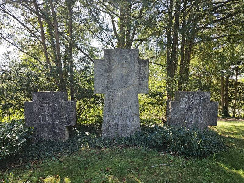 Drei große steinerne Kreuze mit Inschriften stehen in einem von Bäumen umgebenen, grünen Park.