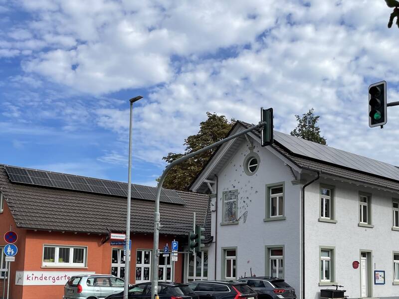 Ein weißes Gebäude mit Solarpanelen und ein rotes Gebäude mit der Aufschrift "Kindergarten", beide unter einem bewölkten Himmel mit einer grünen Ampel im Vordergrund.