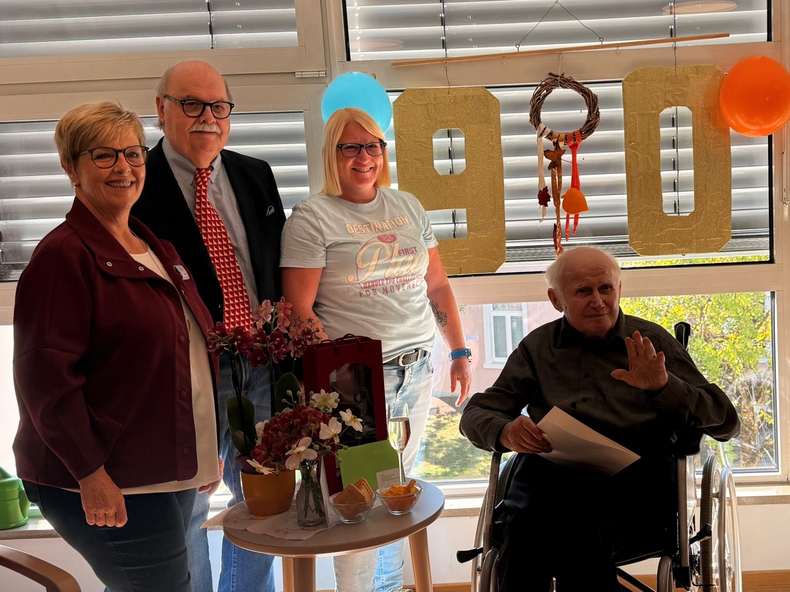 Vier Menschen, darunter eine Person im Rollstuhl, stehen zusammen in einem Raum mit großen goldenen Zahlen "90" im Hintergrund, die auf einen Geburtstag hinweisen.