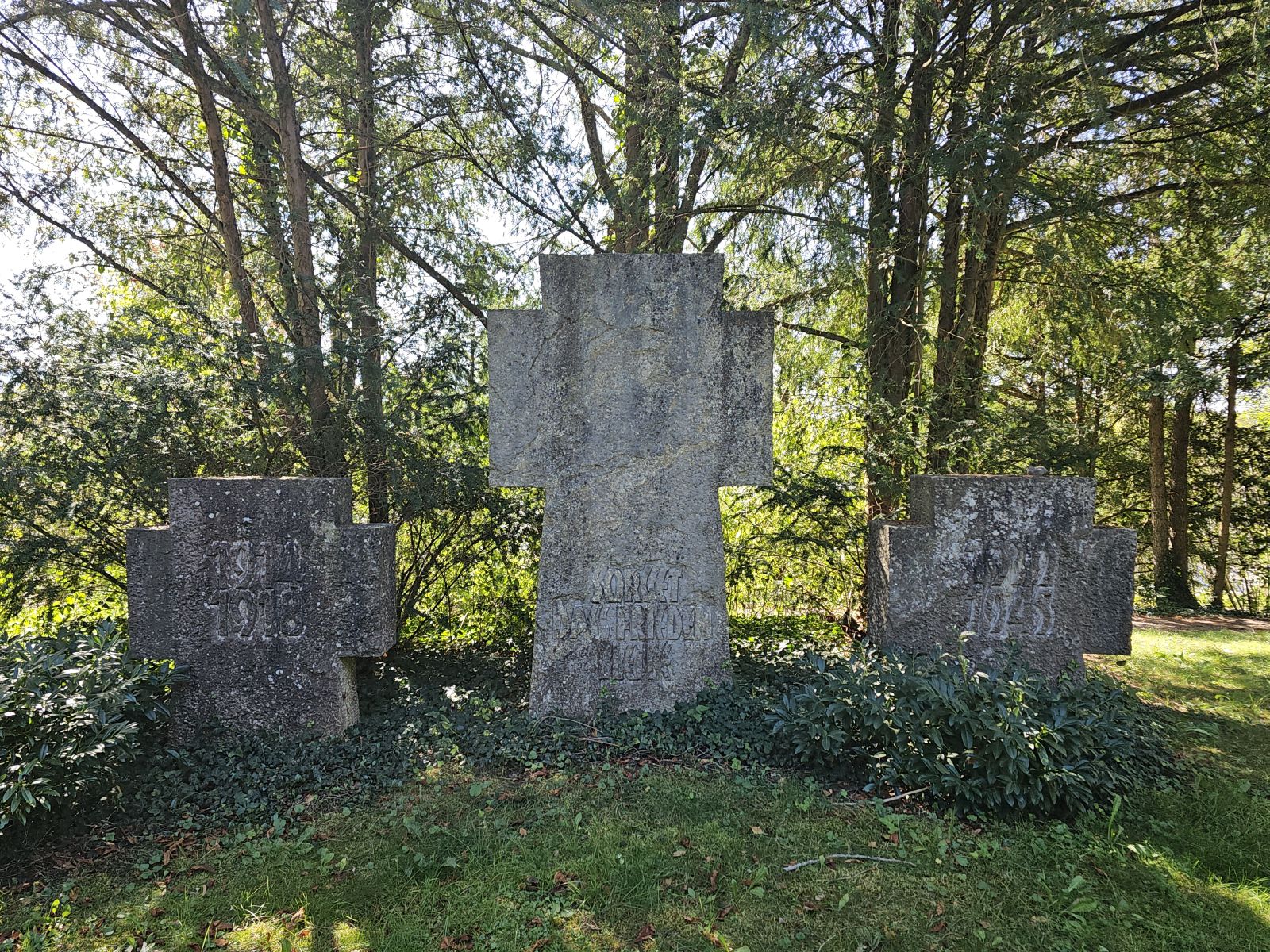 Drei große steinerne Kreuze mit Inschriften stehen in einem von Bäumen umgebenen, grünen Park.